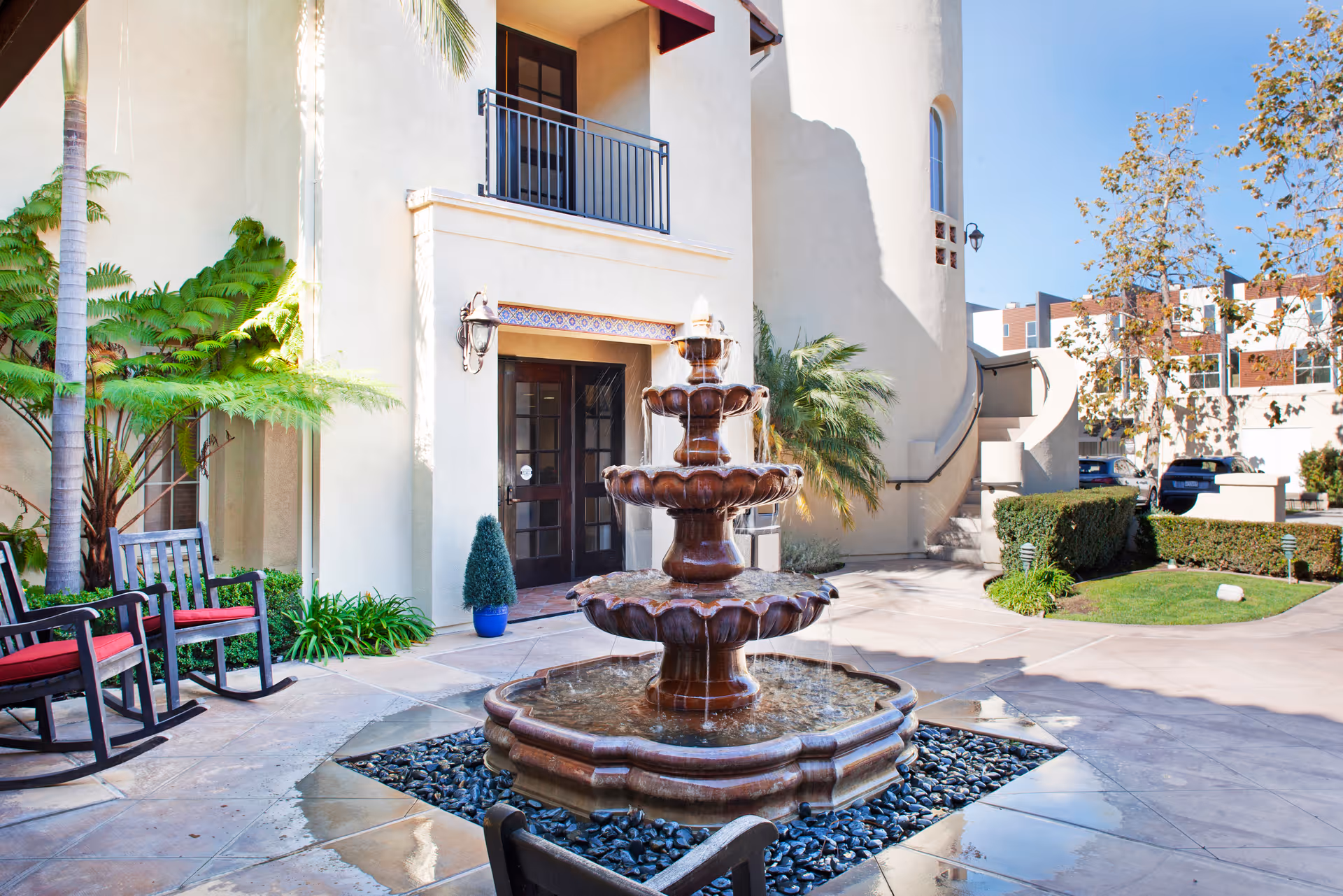 Outdoor courtyard area with a multi-tiered water fountain in the center, surrounded by a tiled patio. There are two rocking chairs with red cushions on the left side near a palm tree and other greenery. The building has beige walls, a balcony with a black railing, and a door with glass panels. Additional plants and trees are visible around the courtyard under a clear blue sky.