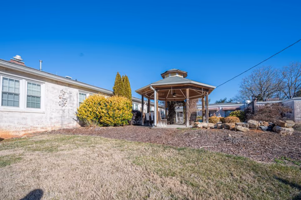 Outdoor view of Harmony House Memory Care featuring a wooden gazebo surrounded by landscaped bushes, rocks, and a grassy area under a clear blue sky.