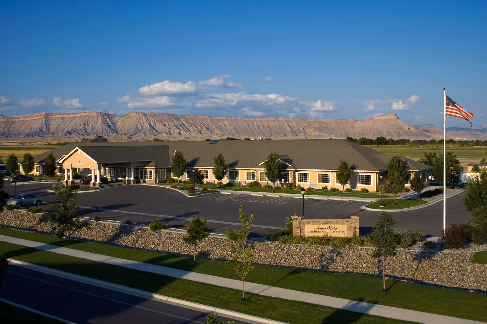 Exterior view of Aspen Ridge Alzheimer’s Special Care Center, a single-story building with a large parking lot, landscaped greenery, and an American flag on a flagpole. In the background, there are expansive flat-topped mountains under a blue sky with scattered clouds.
