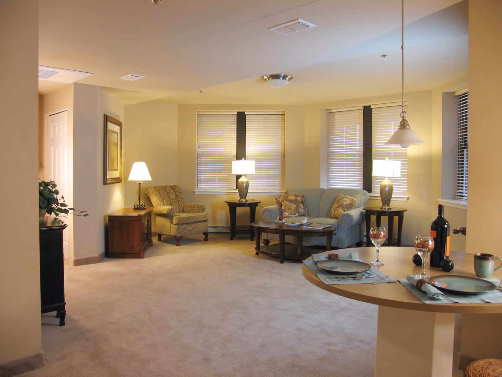 A cozy living room area with beige carpet and walls, featuring a patterned armchair, a light blue sofa with floral cushions, two side tables with lamps, a coffee table with cups and a magazine, and a round dining table set with plates, wine glasses, a bottle of wine, and a mug. The room has several windows with blinds and soft lighting.