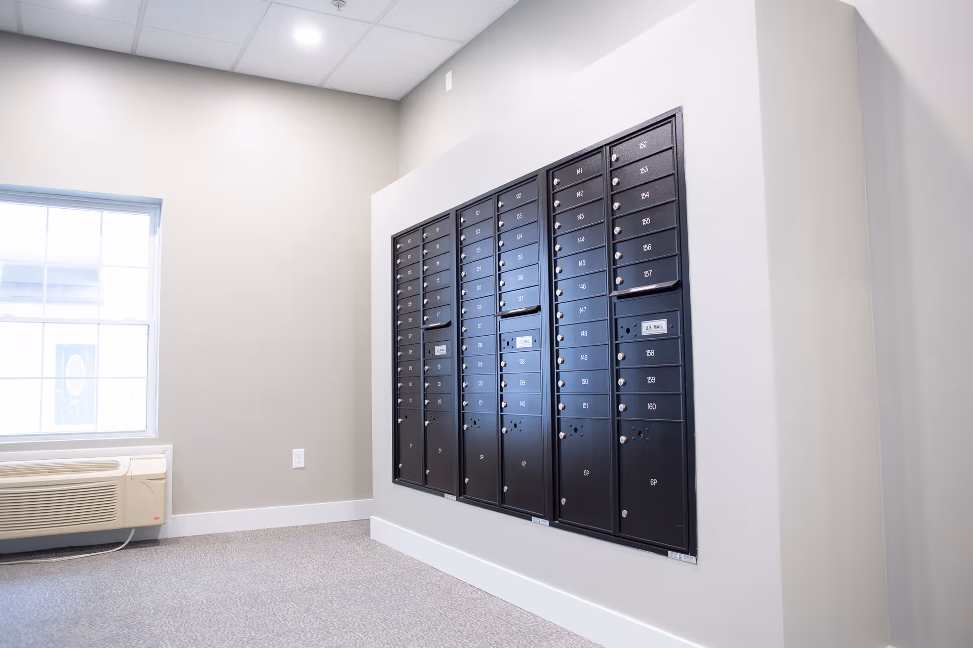 A set of black resident mailboxes mounted on a wall in a bright interior room with a window and an air-conditioning unit.
