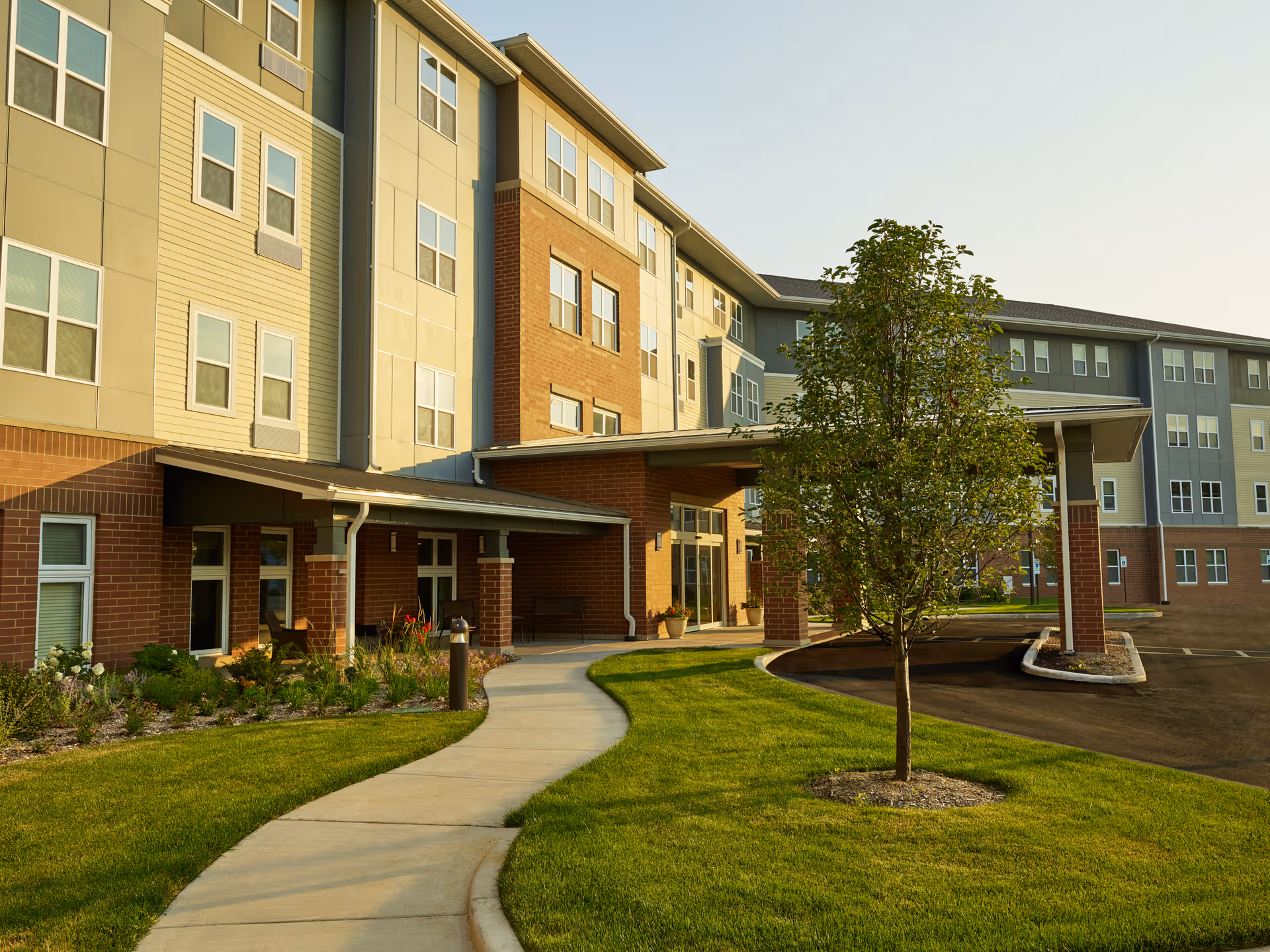 Front entrance of a multi-story senior living building with a covered drop-off, curved walkway, and landscaped lawn with a tree.
