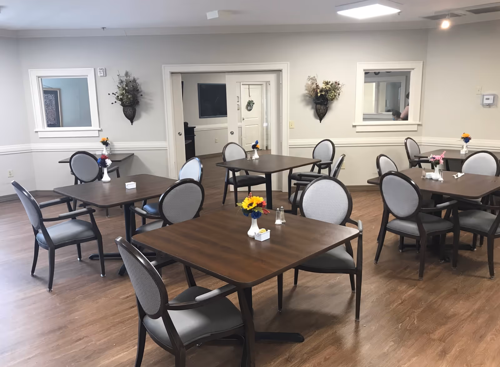 A dining room with several square wooden tables and gray cushioned chairs arranged around them. Each table has a small vase with colorful flowers and salt and pepper shakers. The room has light-colored walls, wooden flooring, two wall-mounted flower arrangements, and windows looking into adjacent rooms.