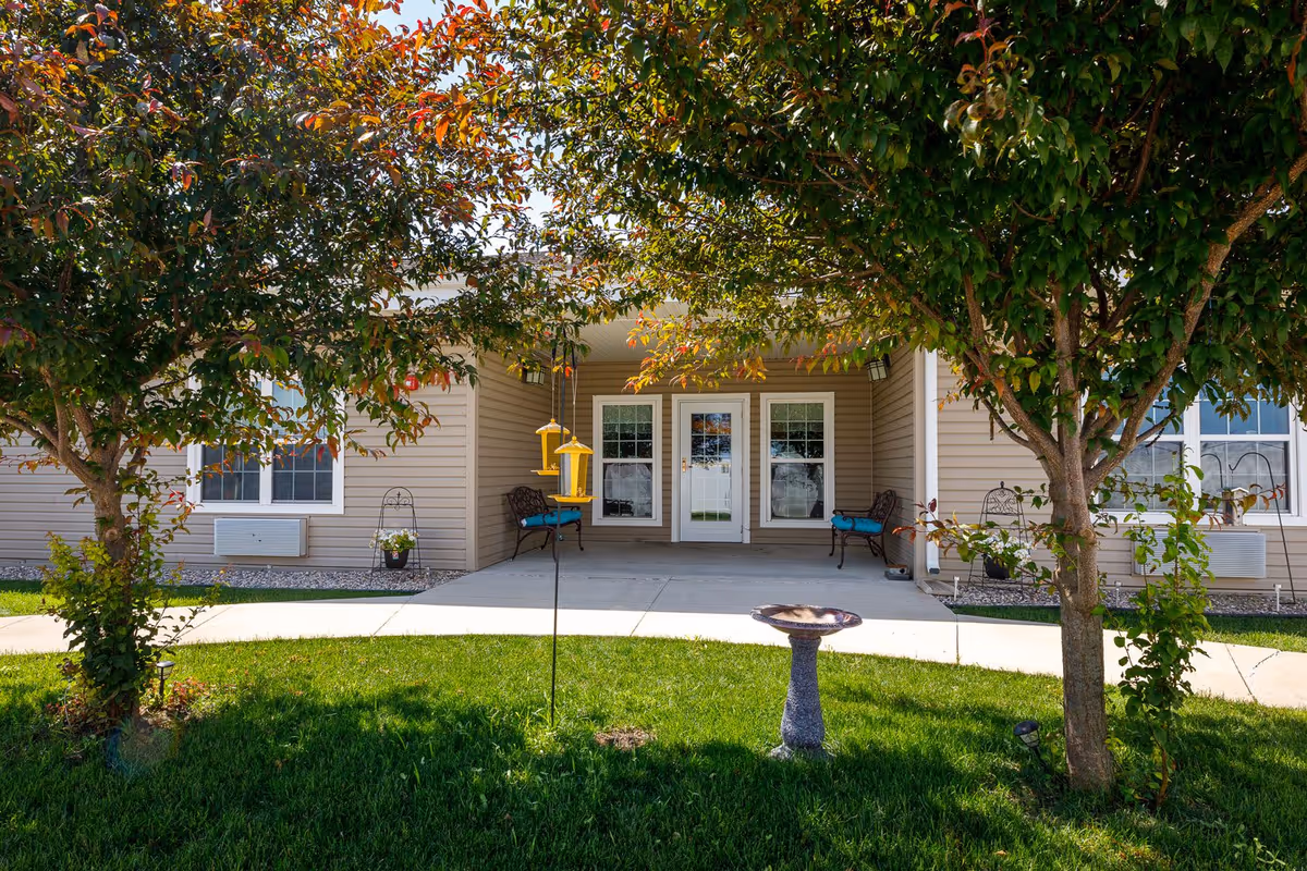 Front entrance of a single-story assisted living facility with a covered porch, benches, trees, a bird feeder, and a birdbath on a grassy lawn.