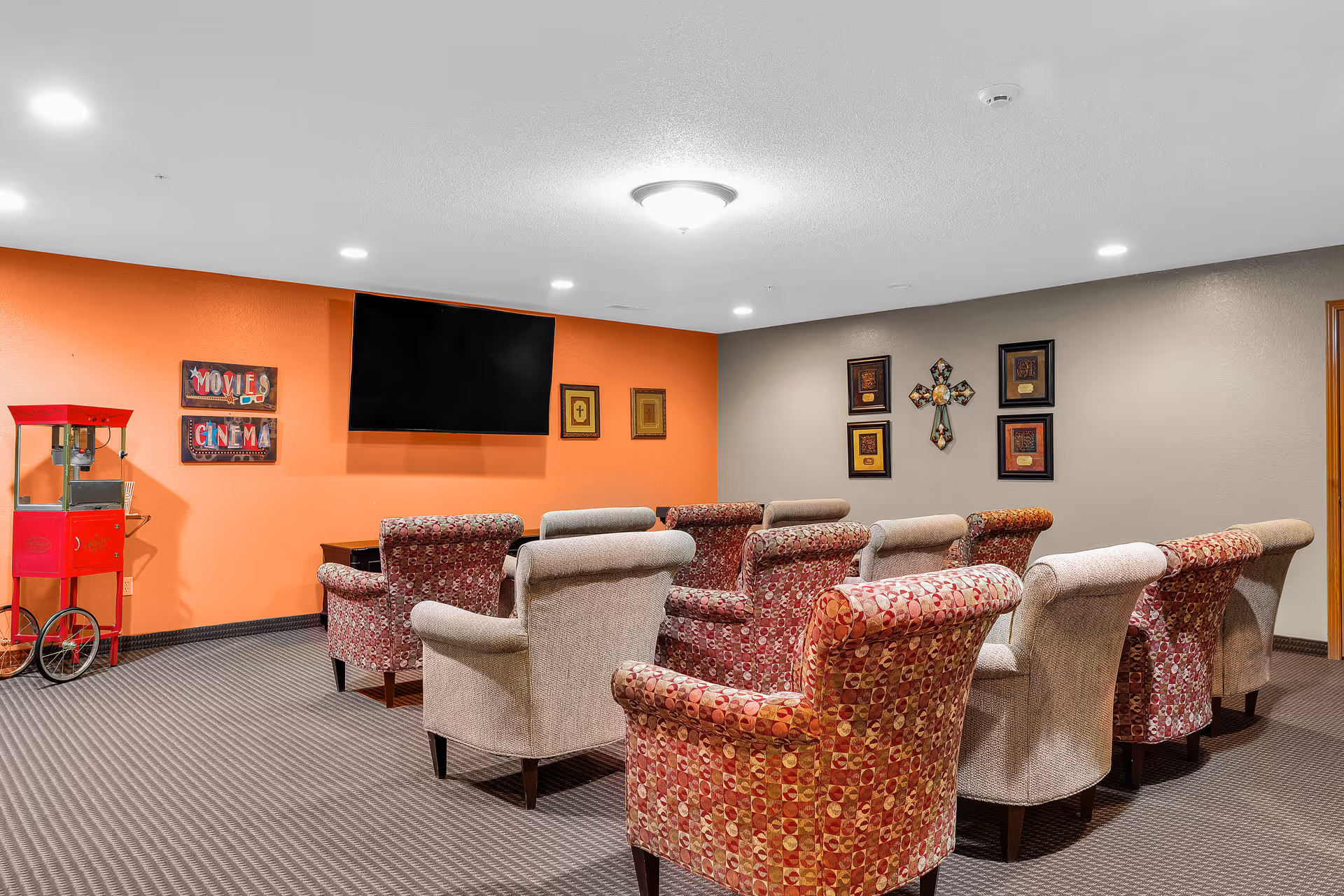 Small screening room with rows of upholstered armchairs facing a wall-mounted TV and a popcorn cart against an orange accent wall.