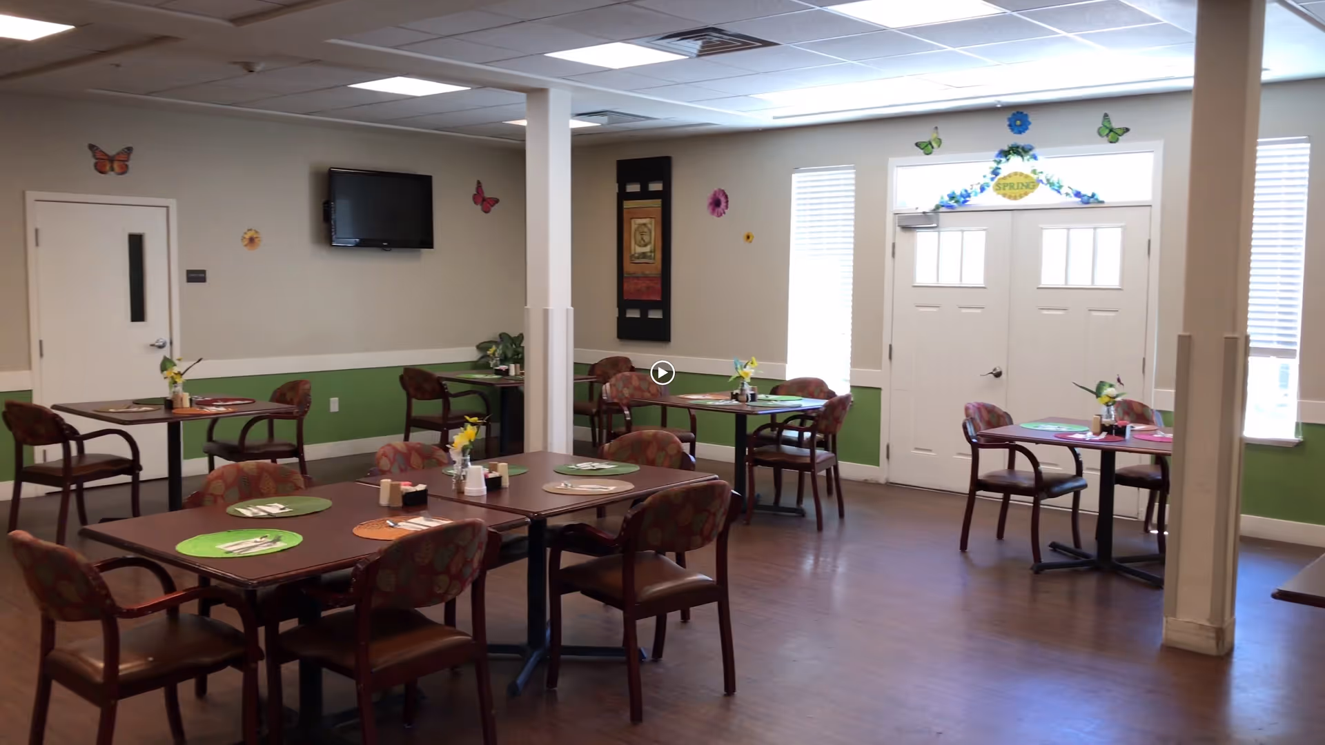 A dining room in an assisted living facility with several tables and chairs arranged neatly. Each table has placemats, utensils, and small flower vases. The walls are decorated with butterfly and flower decals, and there is a television mounted on one wall. Double doors with windows let in natural light.