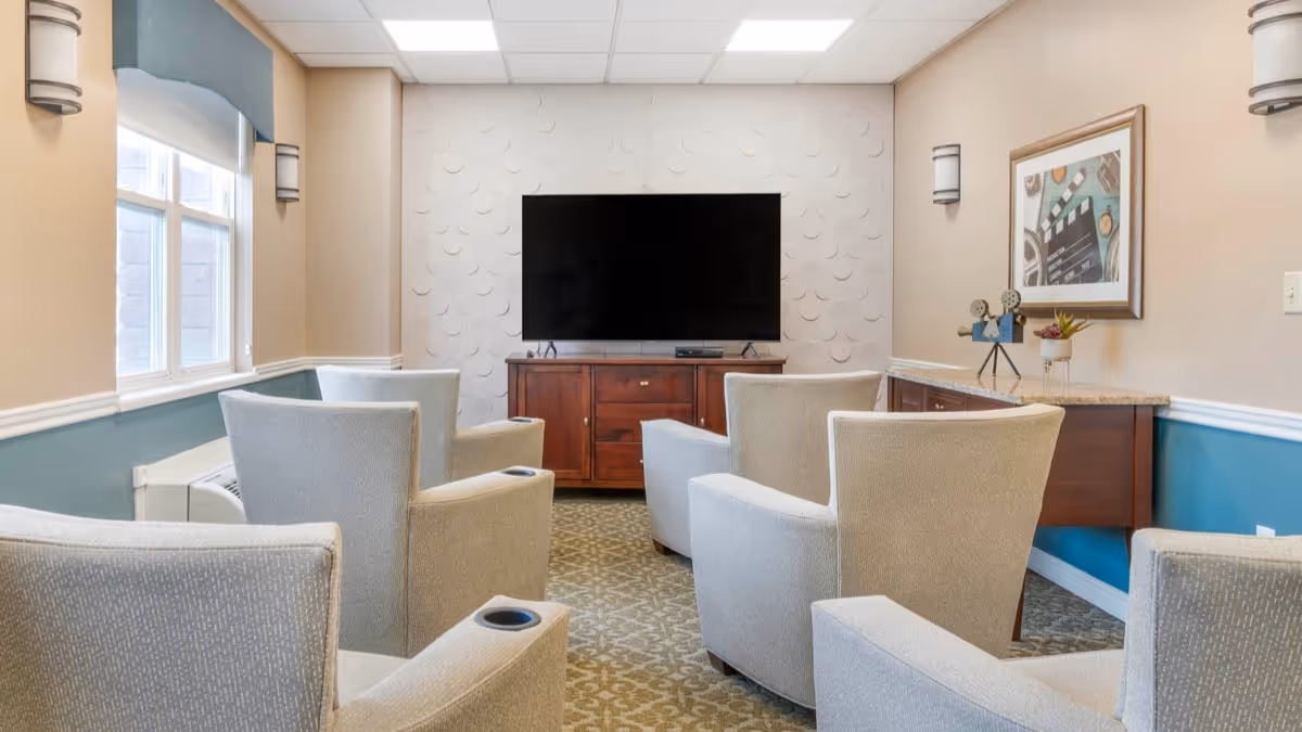 A communal lounge with several upholstered armchairs arranged facing a wall-mounted TV and wooden cabinet.