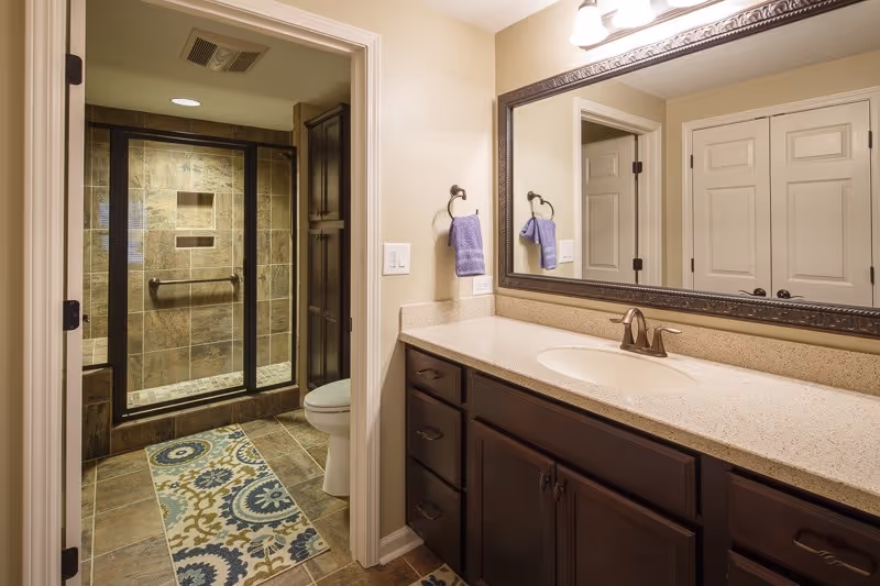 A bathroom with a large vanity featuring a beige countertop, an under-mount sink, and dark wood cabinets. A large framed mirror hangs above the vanity with two purple hand towels on towel rings. In the background, there is a glass-enclosed shower with tiled walls and a built-in niche, a toilet, and a patterned rug on the tiled floor.