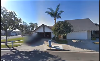 A residential street view showing two houses with garages, a palm tree, and other trees under a clear blue sky.
