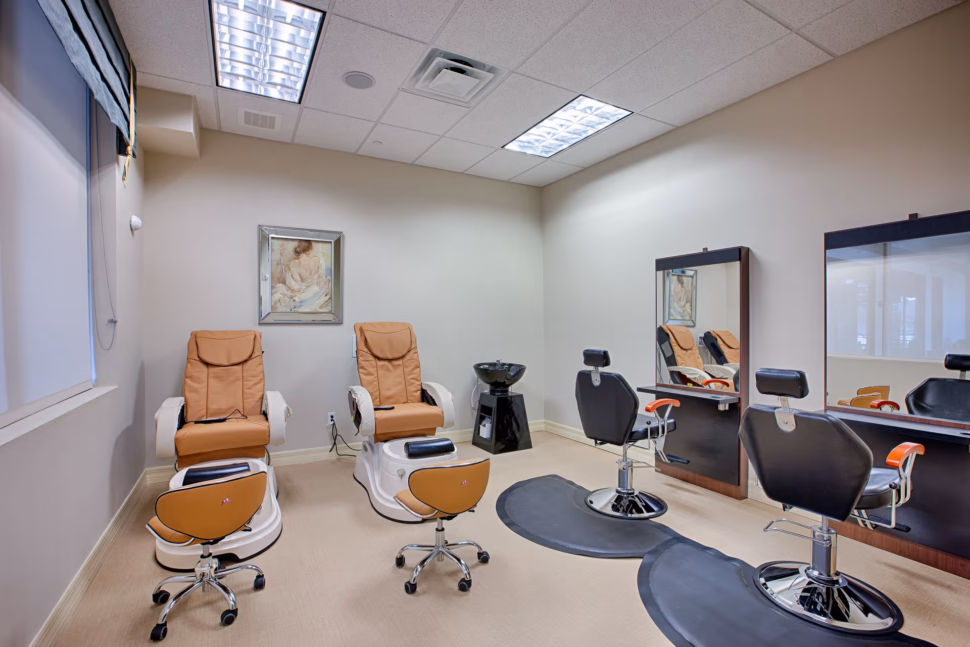 A salon room with two tan pedicure chairs with footrests and two black styling chairs facing mirrors. The room has beige walls, a framed painting on the wall, and overhead fluorescent lighting.