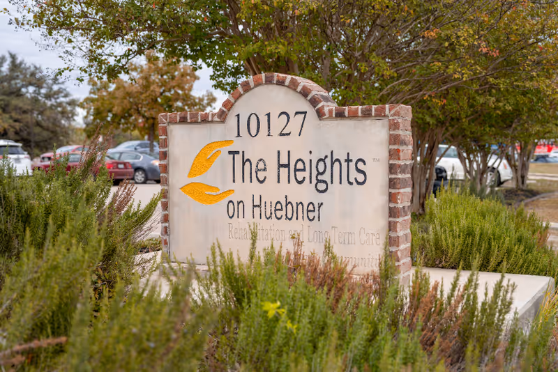A stone and brick sign for The Heights on Huebner, located at 10127, surrounded by greenery and trees with parked cars visible in the background.