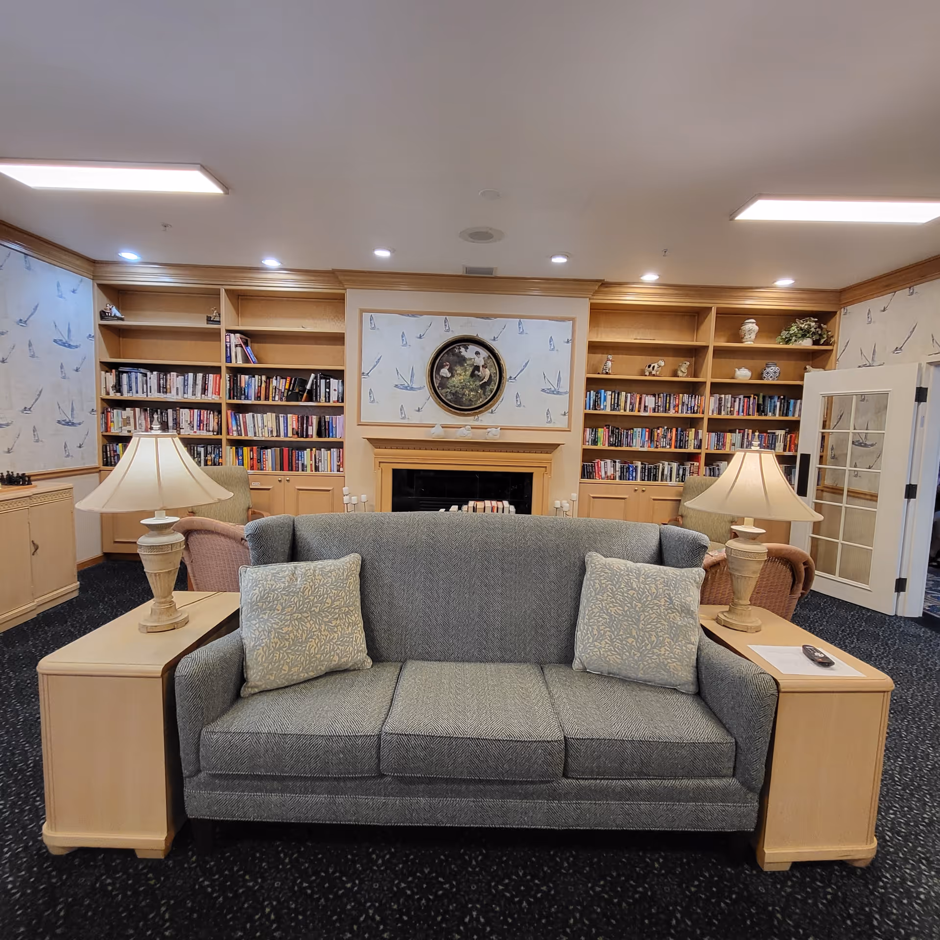 A cozy living room area with a gray upholstered sofa flanked by two light wood side tables, each with a lamp. Behind the sofa is a fireplace with a round framed painting above it, surrounded by built-in wooden bookshelves filled with books and decorative items. The walls have wallpaper with a sailboat pattern, and there are two armchairs partially visible on either side of the fireplace.
