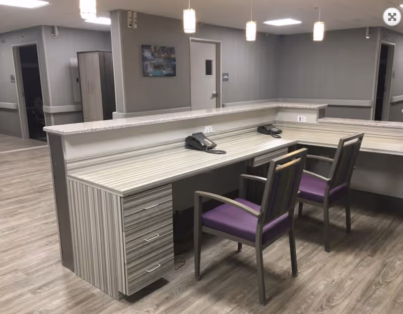 A reception/nurse station inside a senior living facility with a long desk, two telephones and two purple-seated chairs.