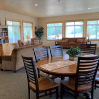 Communal senior living room with a round wooden dining table and chairs in the foreground and sofas, large windows, and lamps in the background.