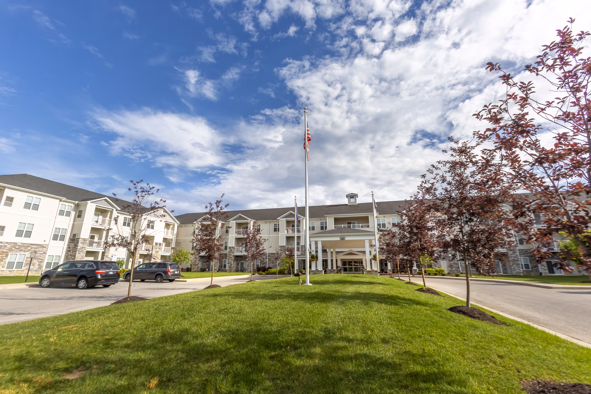 Exterior view of StoryPoint Fort Wayne North, a multi-story senior living facility with a well-maintained lawn, young trees, an American flag on a flagpole, and a partly cloudy blue sky.