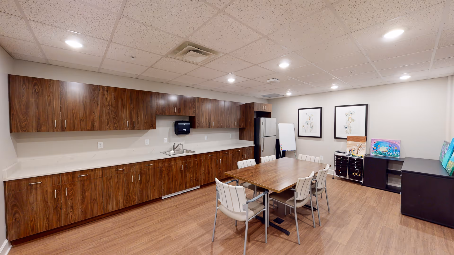 A well-lit room with wood flooring and a long wooden cabinet with a sink and stainless steel refrigerator. In the center, there is a wooden table surrounded by six cushioned chairs. The walls are decorated with two framed floral artworks, and there are colorful paintings and art supplies on black storage units along the right wall. The ceiling has recessed lighting and a drop ceiling with ventilation.