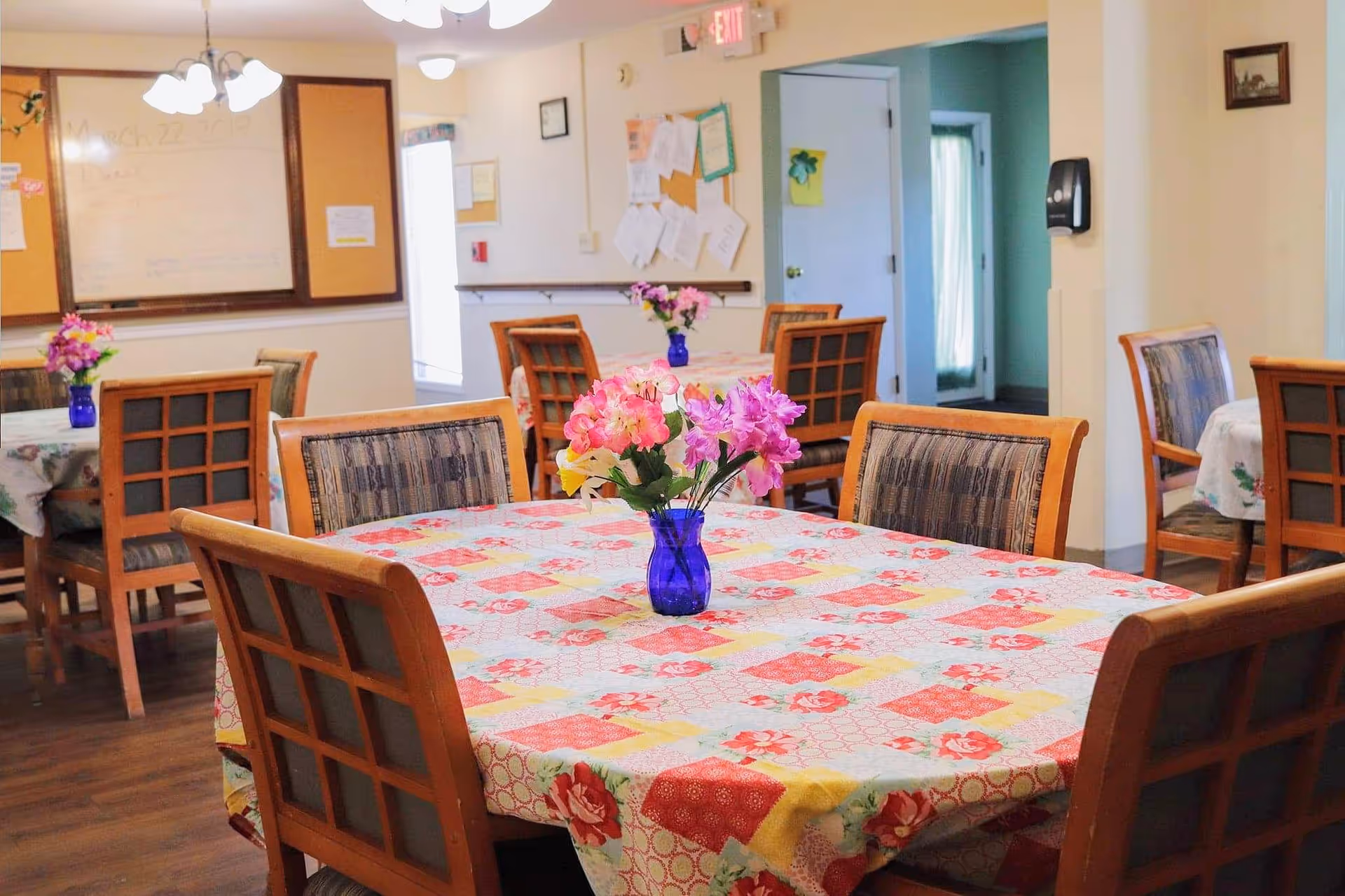 A dining room with several wooden tables covered with floral-patterned tablecloths. Each table has a blue vase with pink and purple flowers. Wooden chairs with cushioned seats surround the tables. The room has light-colored walls, a bulletin board, and a whiteboard in the background.