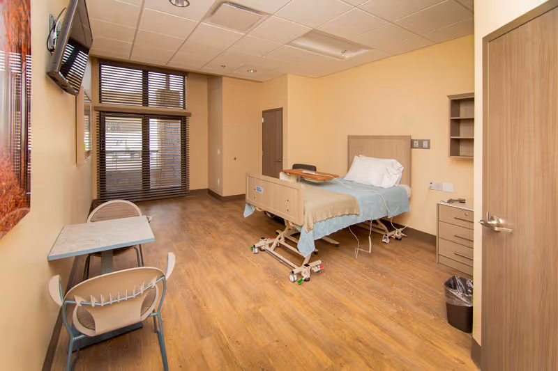 A bright private senior-care bedroom with an adjustable hospital-style bed, small table and chairs, wall-mounted TV, and wood-look flooring.