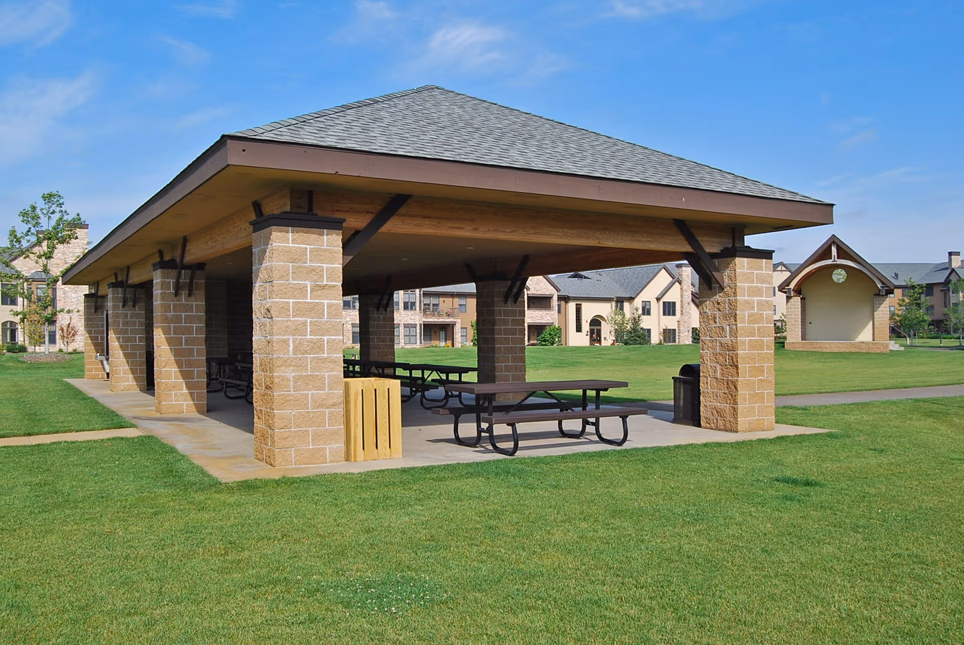 A covered outdoor pavilion with picnic tables underneath, situated on a well-maintained grassy area with residential buildings in the background under a clear blue sky.