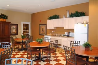 A dining area with several round wooden tables and metal chairs arranged on a checkered floor. The background features a kitchenette with white cabinets, a refrigerator, a microwave, and decorative plants on top of the cabinets. There are framed pictures on the walls and a wooden cabinet on the left side.
