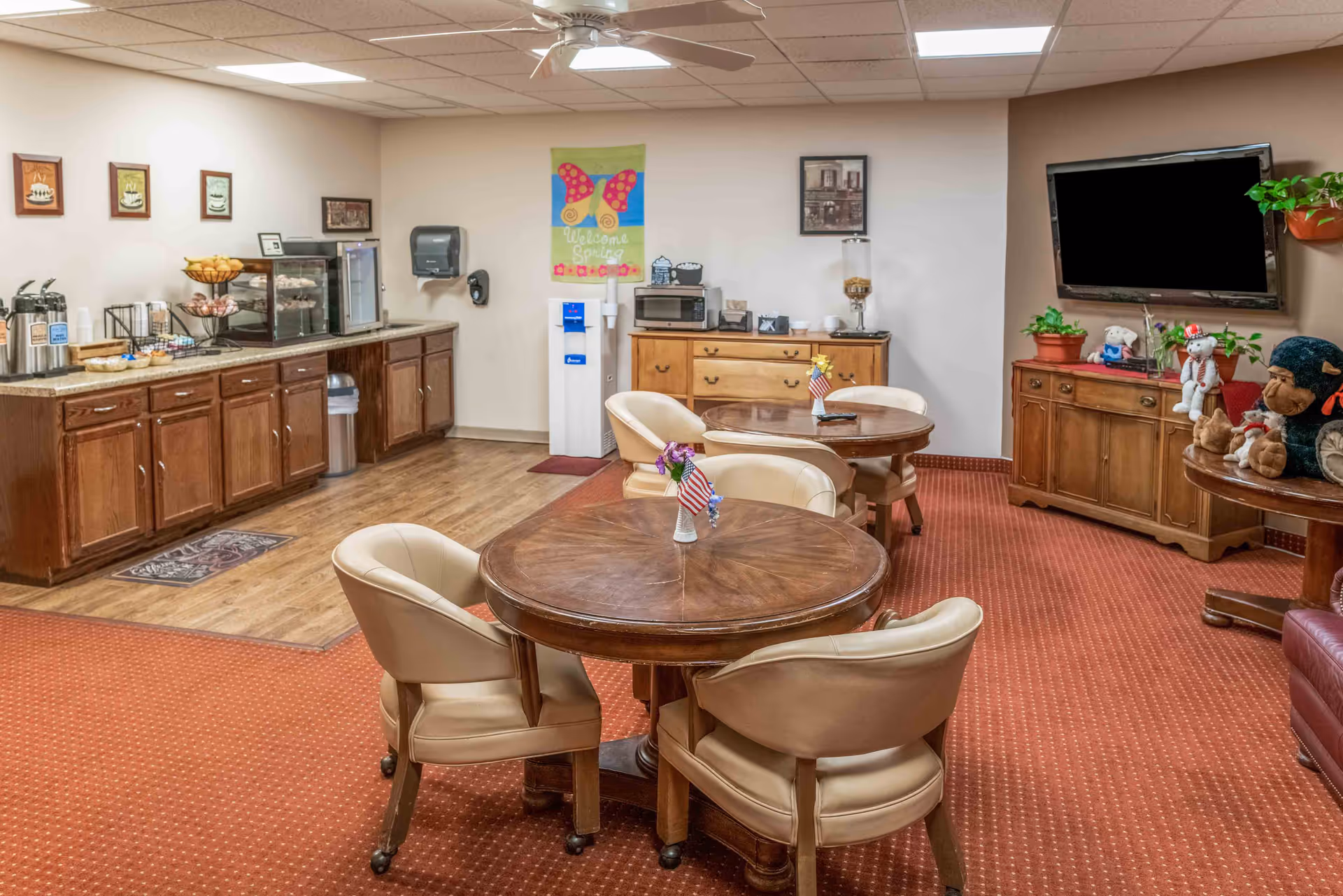 Communal lounge/dining room with round wooden tables and chairs, a snack counter along the wall, TV, and cabinets.