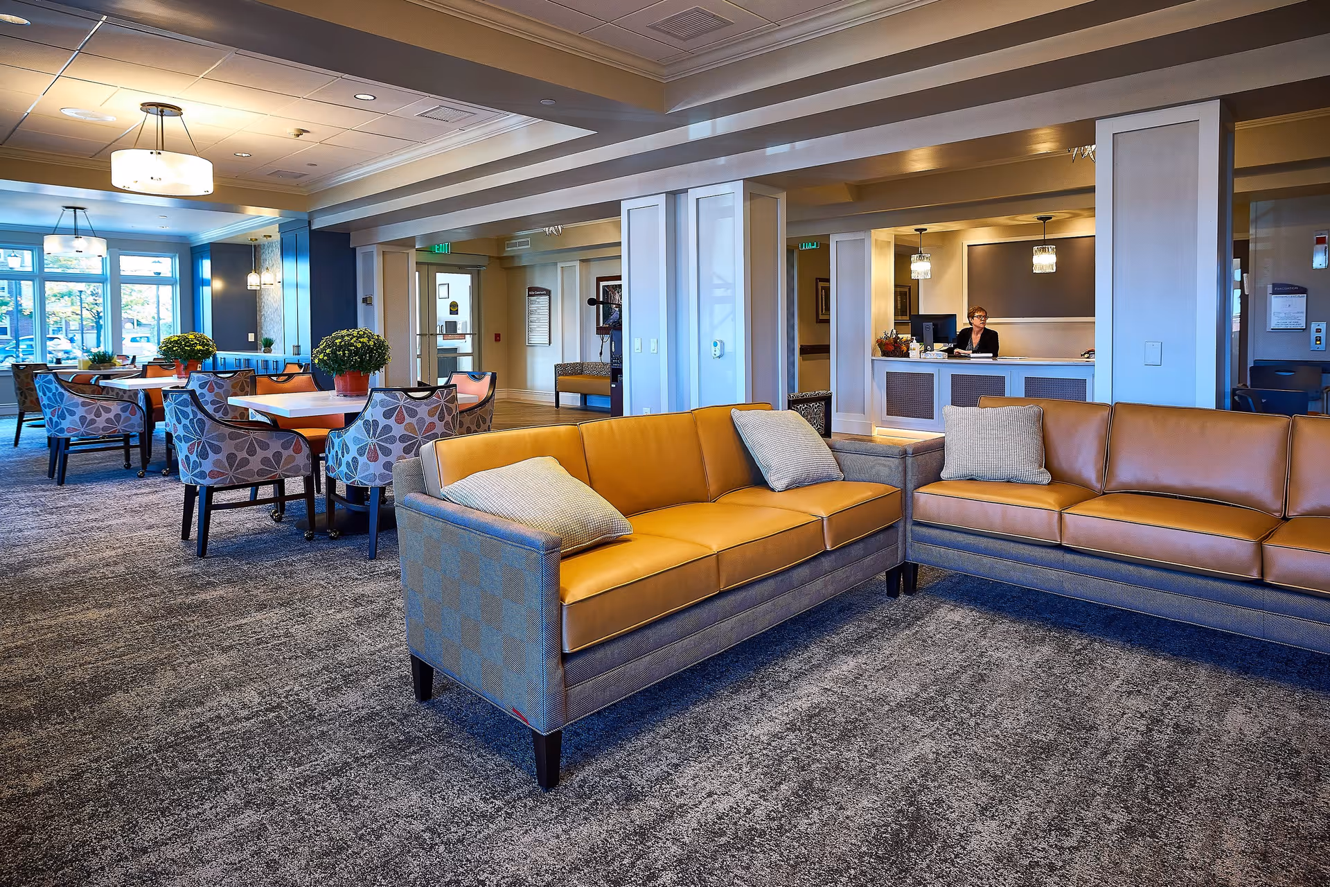 A spacious and well-lit common area in a senior living facility featuring two tan leather sofas with cushions, several round tables with patterned chairs, potted plants on the tables, and a reception desk with a staff member seated behind it. Large windows allow natural light to fill the room.