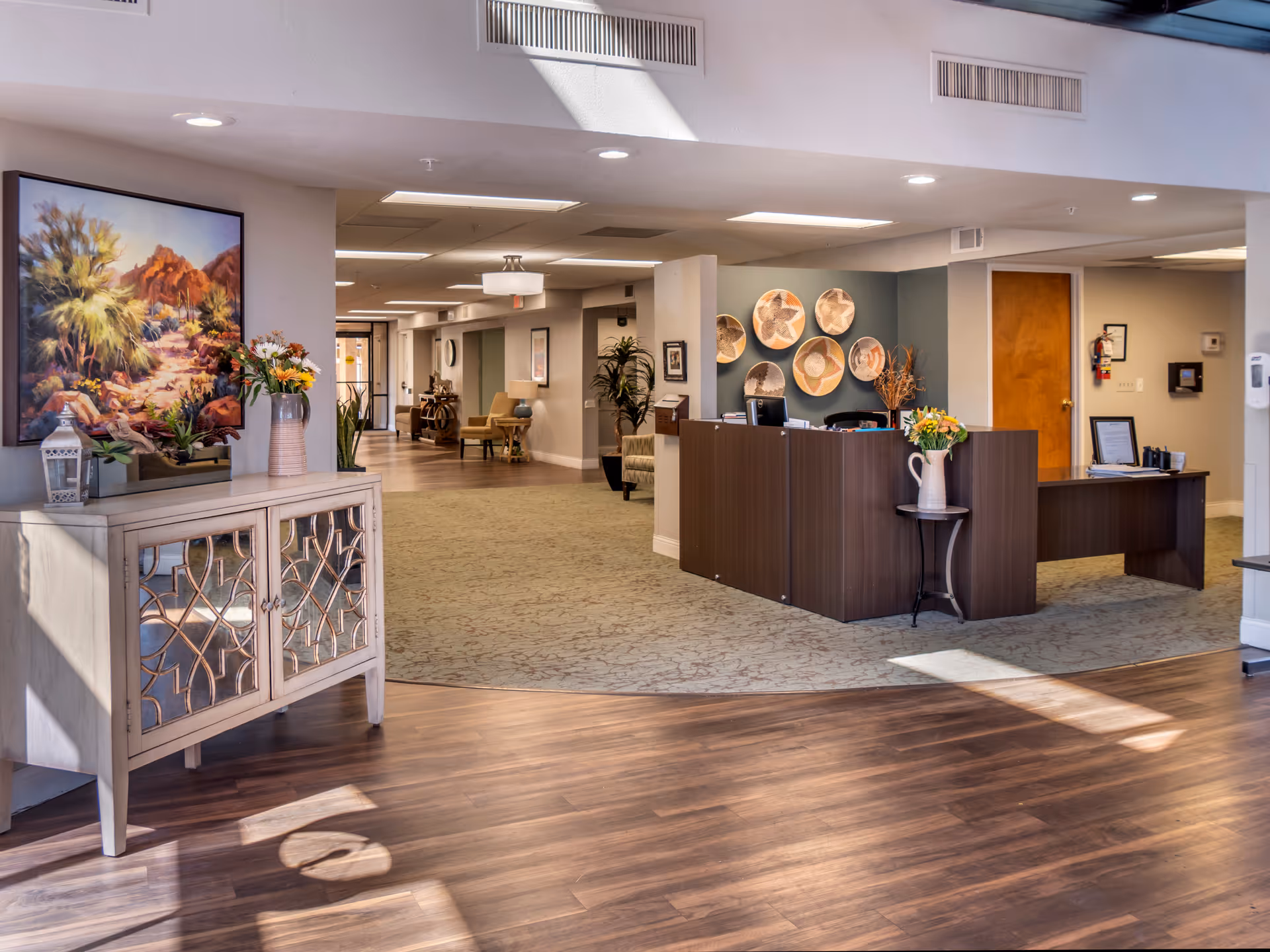 A bright and welcoming reception area in a senior living facility with a wooden reception desk adorned with a vase of flowers. The room features a combination of wood and carpet flooring, decorative wall art including a large painting of a desert landscape and woven baskets, and several plants. The hallway in the background has seating areas with chairs and tables.
