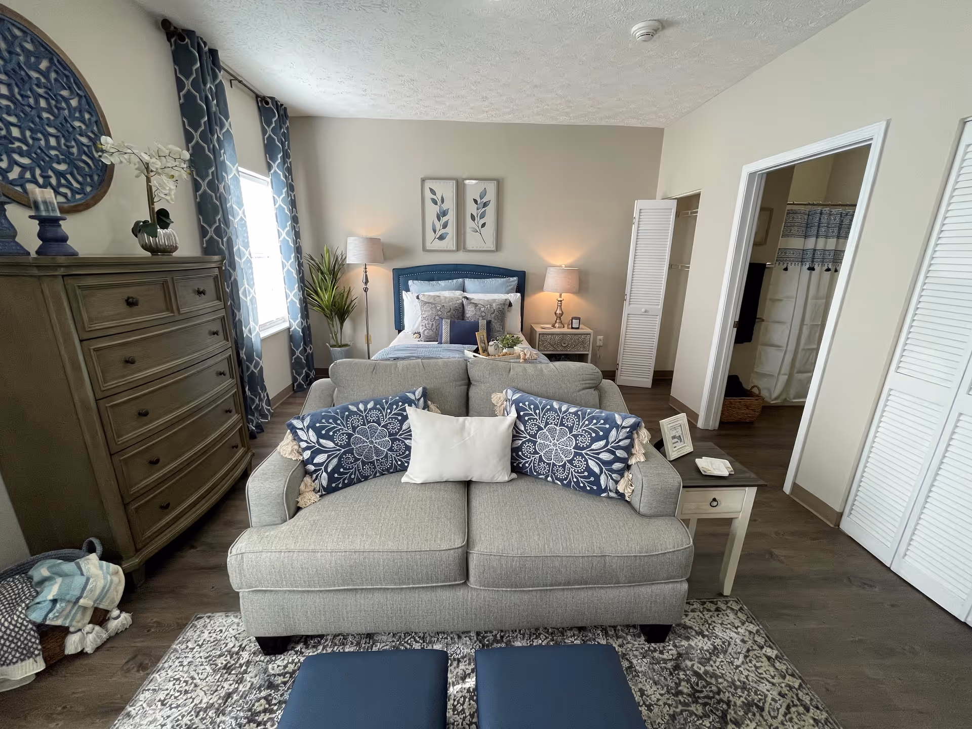 A decorated bedroom suite with a loveseat facing a bed, dresser, nightstands, and an open closet leading to a bathroom.