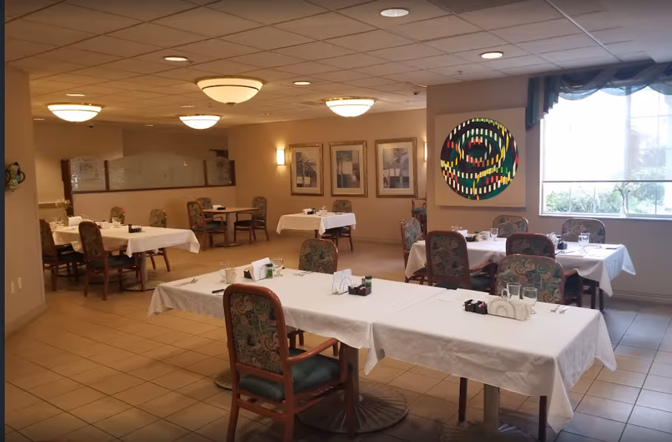 Dining room with multiple tables set with white tablecloths and patterned chairs, wall artwork and large windows.
