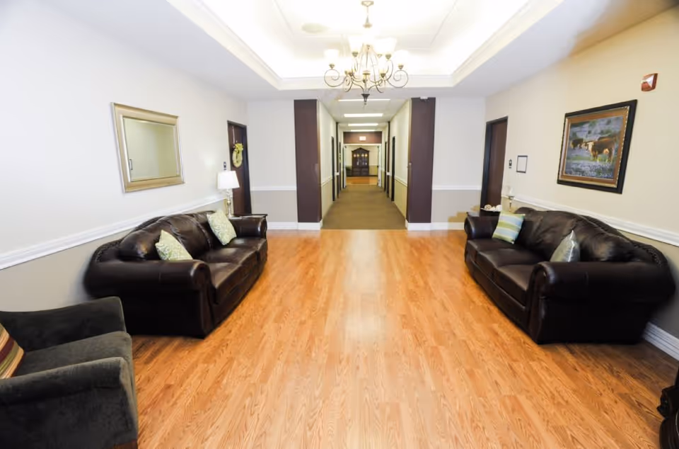 A bright assisted-living lounge with two leather sofas, a chandelier, and a central hallway leading to rooms.