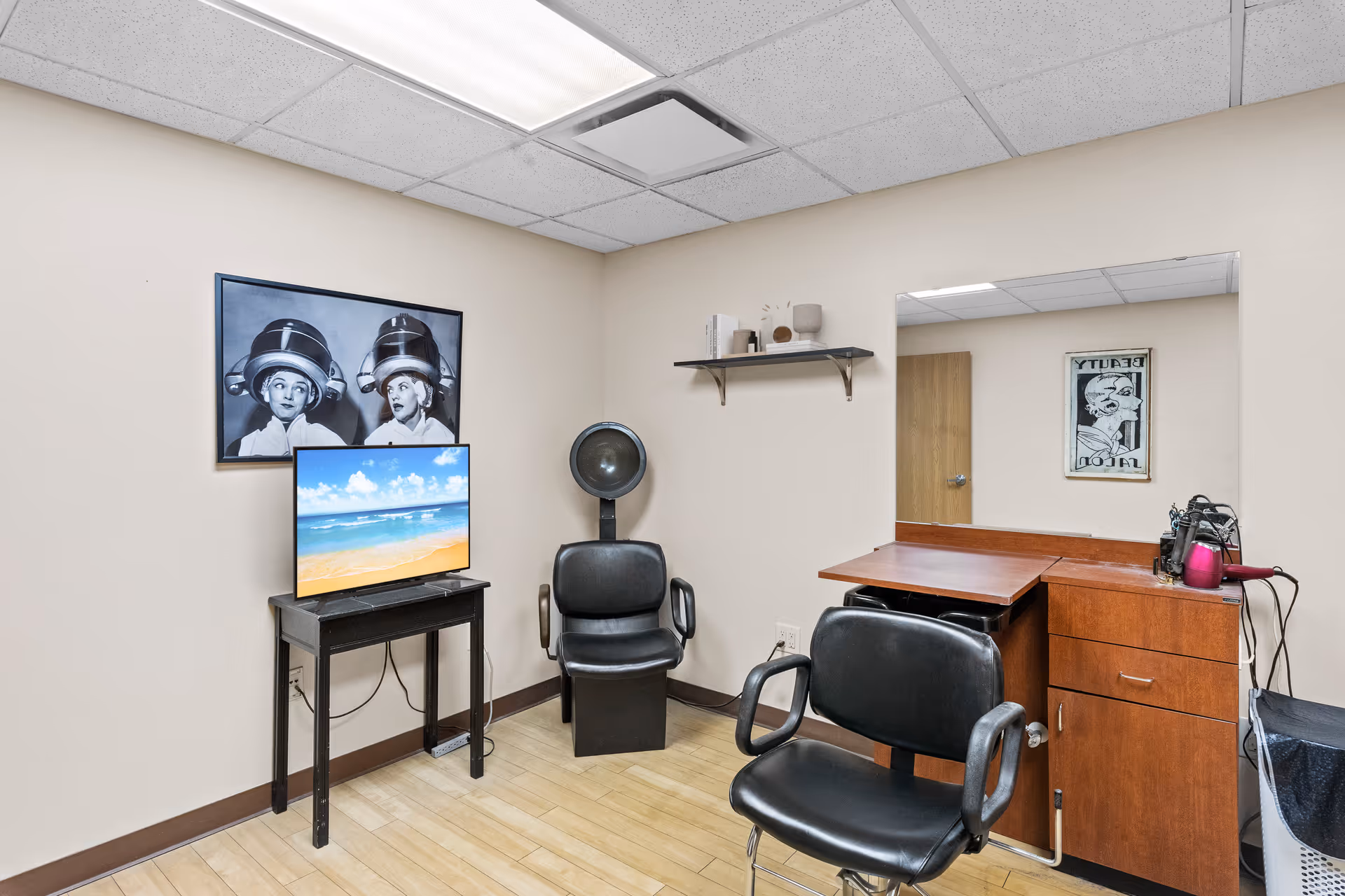 A small salon room with two black salon chairs, a hair dryer hood, a wooden cabinet with a mirror, a hair dryer, and a TV on a small black table displaying a beach scene. The walls are beige with a black and white photo of two women under hair dryers and a shelf with decorative items.