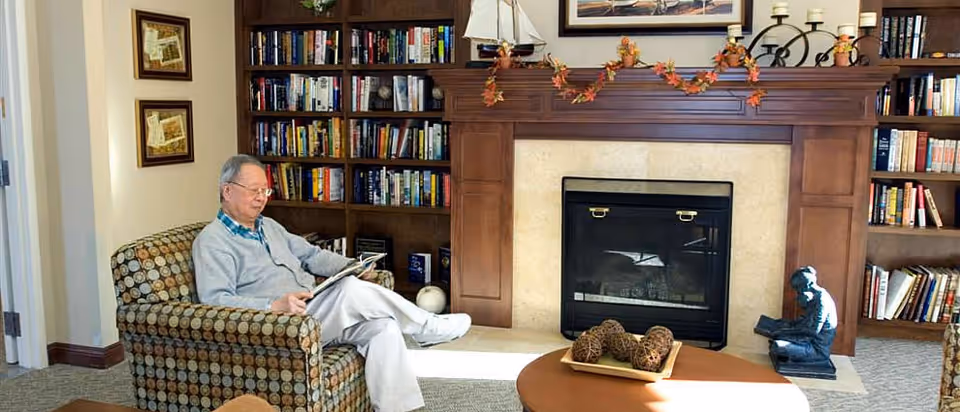 An elderly man reads in an armchair in a cozy common room with bookshelves and a fireplace.