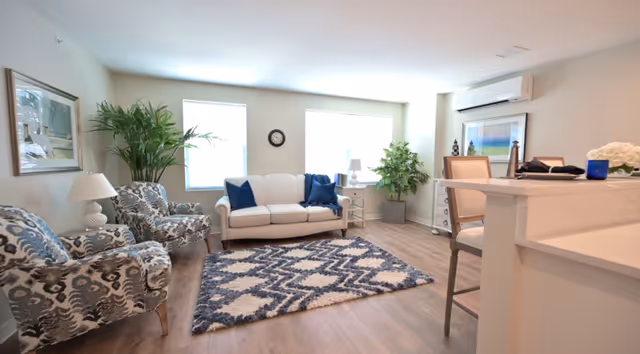 A bright and cozy living room with a white sofa adorned with blue pillows, two patterned armchairs, a blue and white geometric rug, two large windows letting in natural light, potted plants, a wall clock, and a white countertop with chairs on the right side.