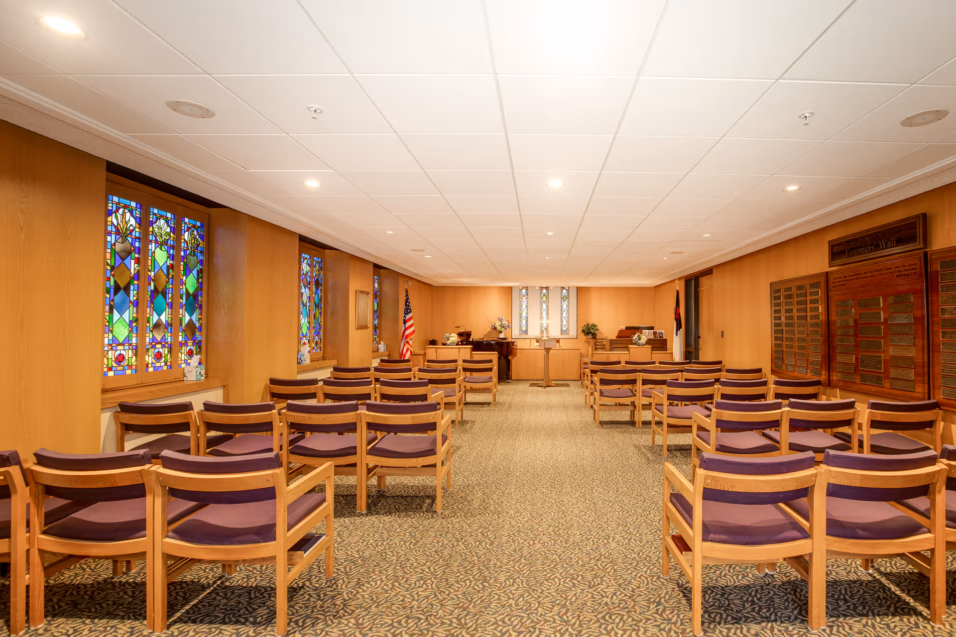 Interior view of a chapel or meeting room with wooden walls, stained glass windows on the left, rows of wooden chairs with purple cushions arranged facing a podium and piano at the front. An American flag and a Christian flag are positioned near the front, and a wooden plaque wall is visible on the right side.