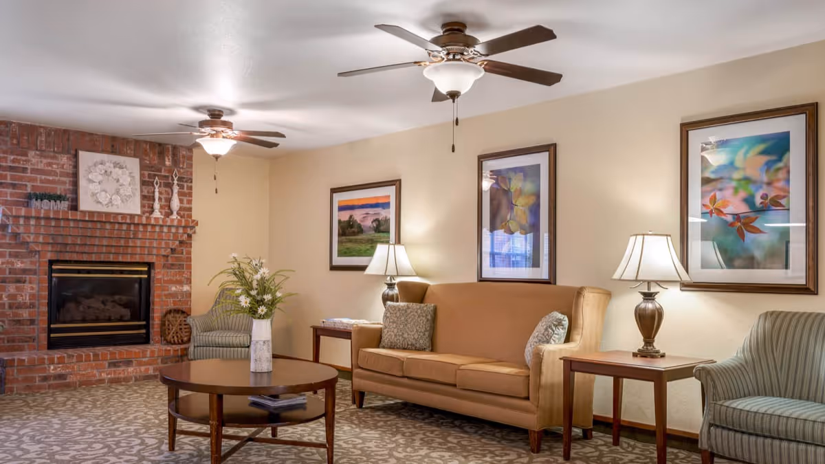 A cozy living room with a brick fireplace on the left, a round wooden coffee table with a vase of flowers in the center, a tan sofa with two pillows, two side tables each with a lamp, and two striped armchairs. The walls are decorated with three framed pictures, and two ceiling fans with lights are mounted on the ceiling.