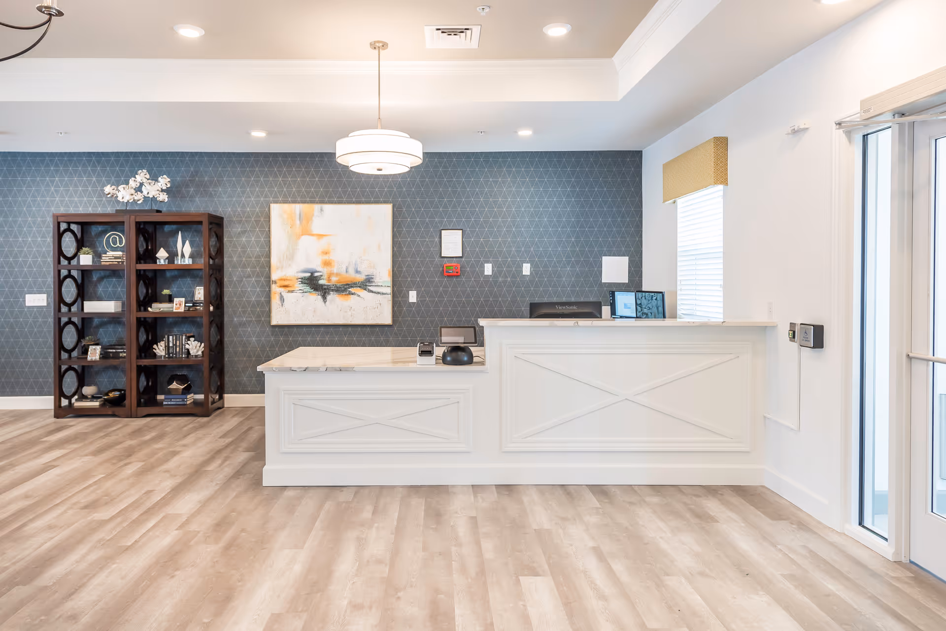 A bright and modern reception area with a white front desk, a computer monitor, and a card reader. Behind the desk is a blue wall with a geometric pattern, a large abstract painting, and a dark wooden bookshelf displaying decorative items. The floor is light wood, and there is a window with a yellow valance to the right, next to glass doors.