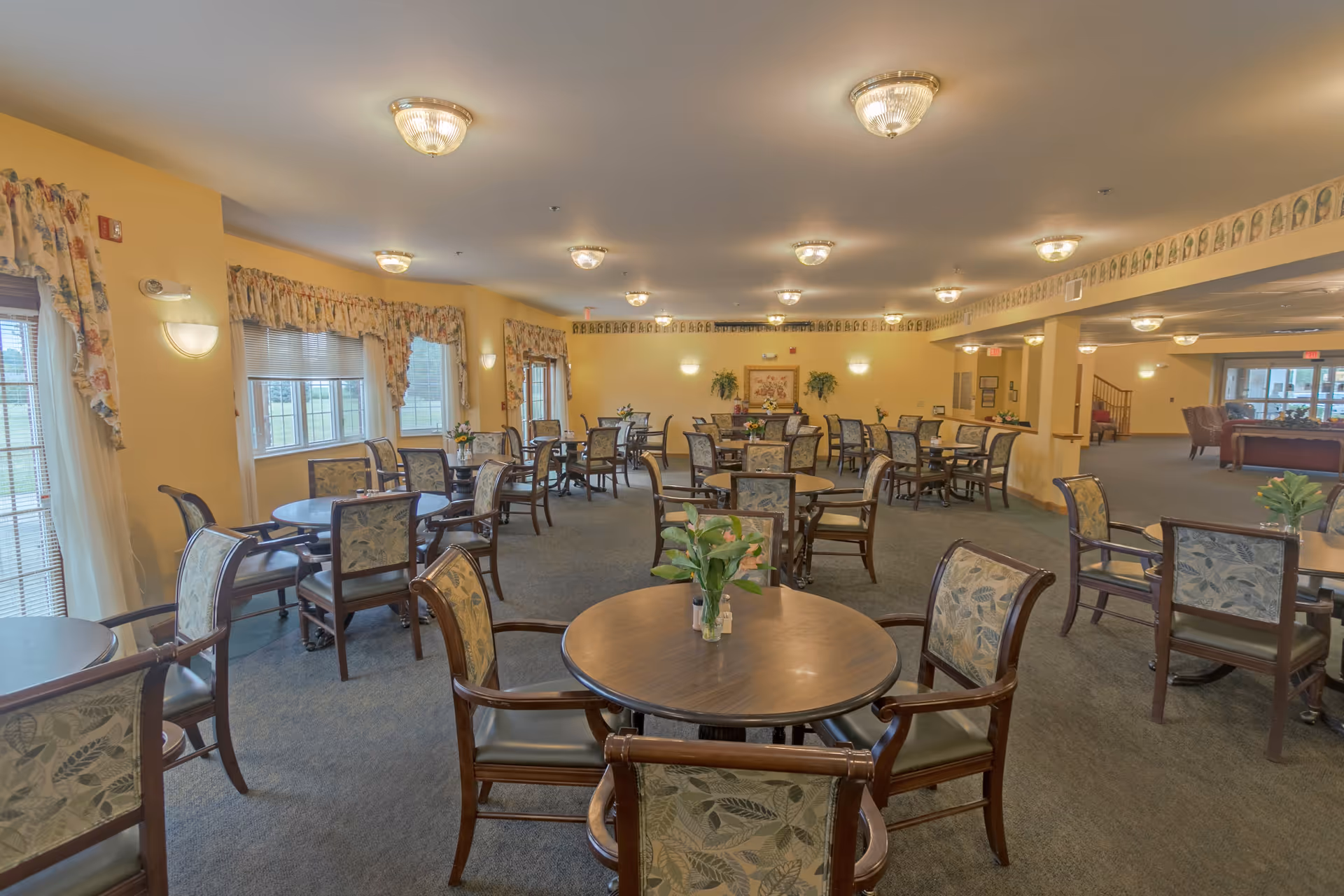 A spacious dining room in a senior living facility with multiple round wooden tables and cushioned chairs arranged neatly. The room has yellow walls, several windows with floral curtains, and ceiling lights providing warm illumination. There are small flower arrangements on the tables and some framed artwork and plants on the far wall.
