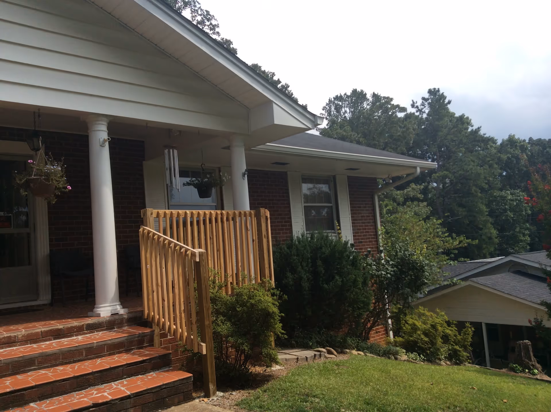 Front porch of a brick building with white columns, a tiled staircase with a wooden handrail, hanging flower pots, and surrounding greenery including bushes and trees.