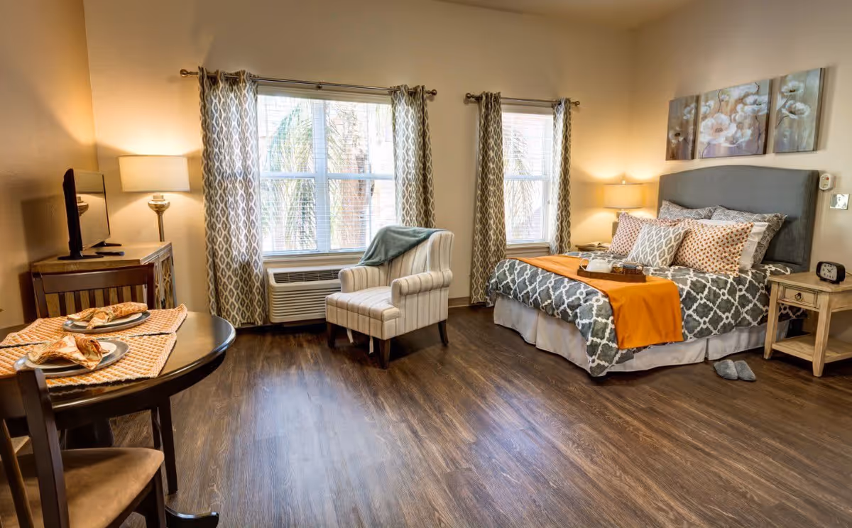 A well-lit bedroom with a large bed covered in patterned bedding and an orange throw blanket. There are multiple pillows on the bed and a pair of slippers on the floor nearby. Next to the bed is a nightstand with a lamp and a clock. A comfortable armchair is positioned near two windows with patterned curtains. A small dining table with two chairs is set with plates and napkins. The room has wooden flooring and warm lighting from lamps.