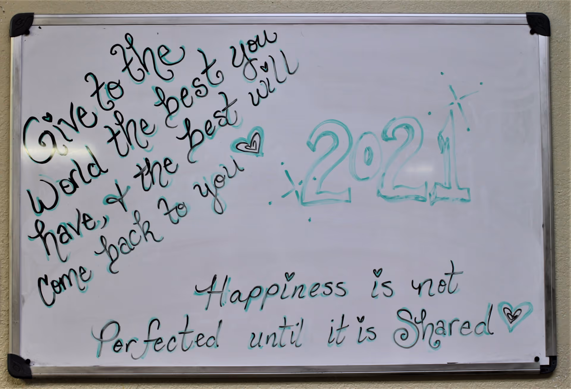 A whiteboard with handwritten inspirational messages in black and teal marker. The messages read: 'Give to the world the best you have, & the best will come back to you' with a heart symbol, '2021' decorated with sparkles, and 'Happiness is not perfected until it is shared' with a heart symbol.