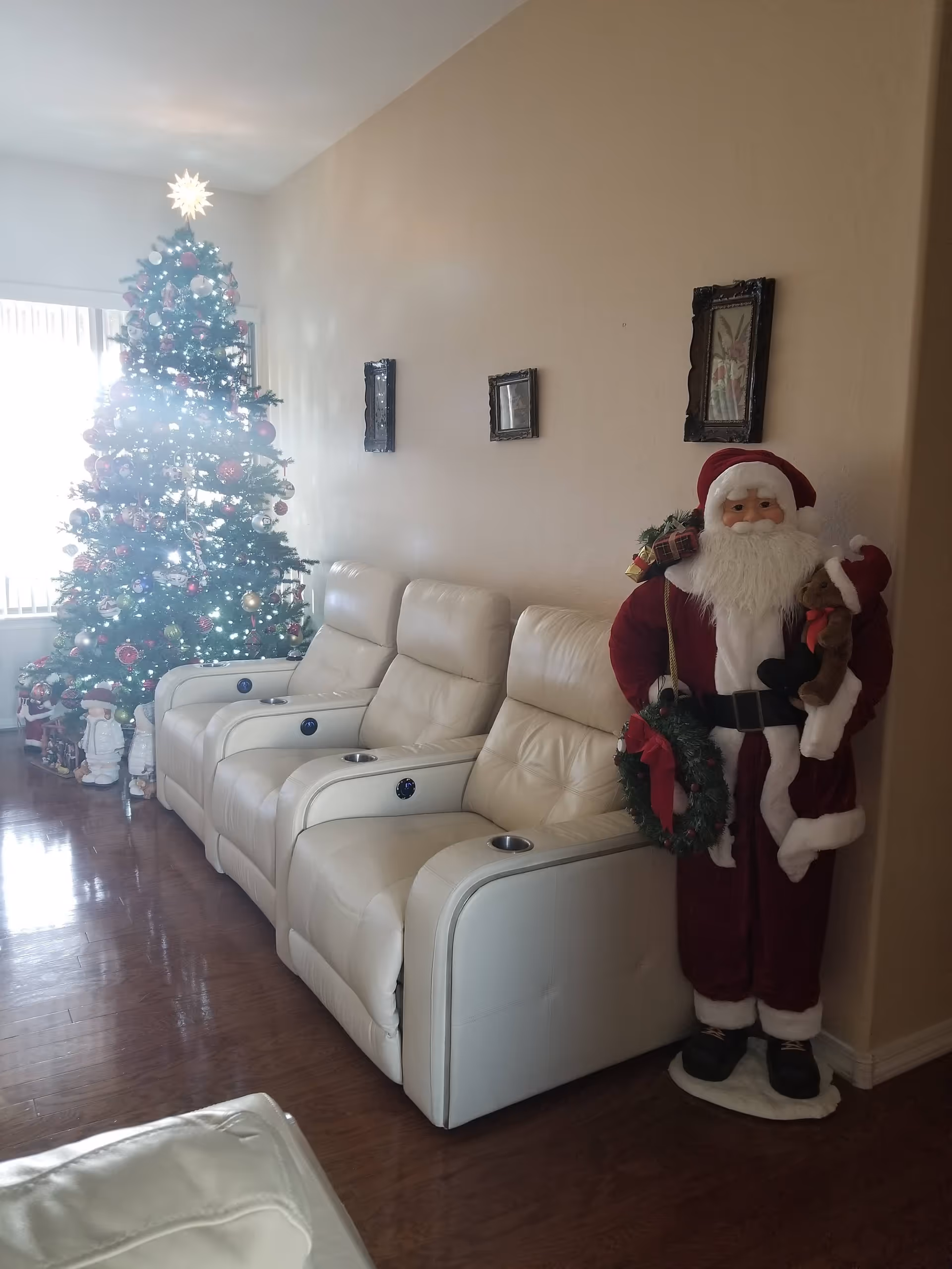 A living room decorated for Christmas with a tall, lit Christmas tree adorned with ornaments and a star on top. Next to the tree are three white leather recliner chairs with cup holders. To the right of the chairs stands a large Santa Claus figure holding a teddy bear and a wreath. The room has wooden flooring and beige walls with three small framed pictures hanging above the chairs.