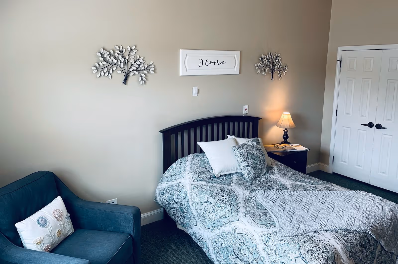 A cozy bedroom with a bed featuring a patterned blue and white comforter and pillows. Above the bed is a wall decoration with the word 'Home' and two metal tree wall art pieces on either side. To the right of the bed is a nightstand with a lit lamp and some items on it. To the left is a blue armchair with a decorative pillow. There is a white double door closet on the right side of the room.