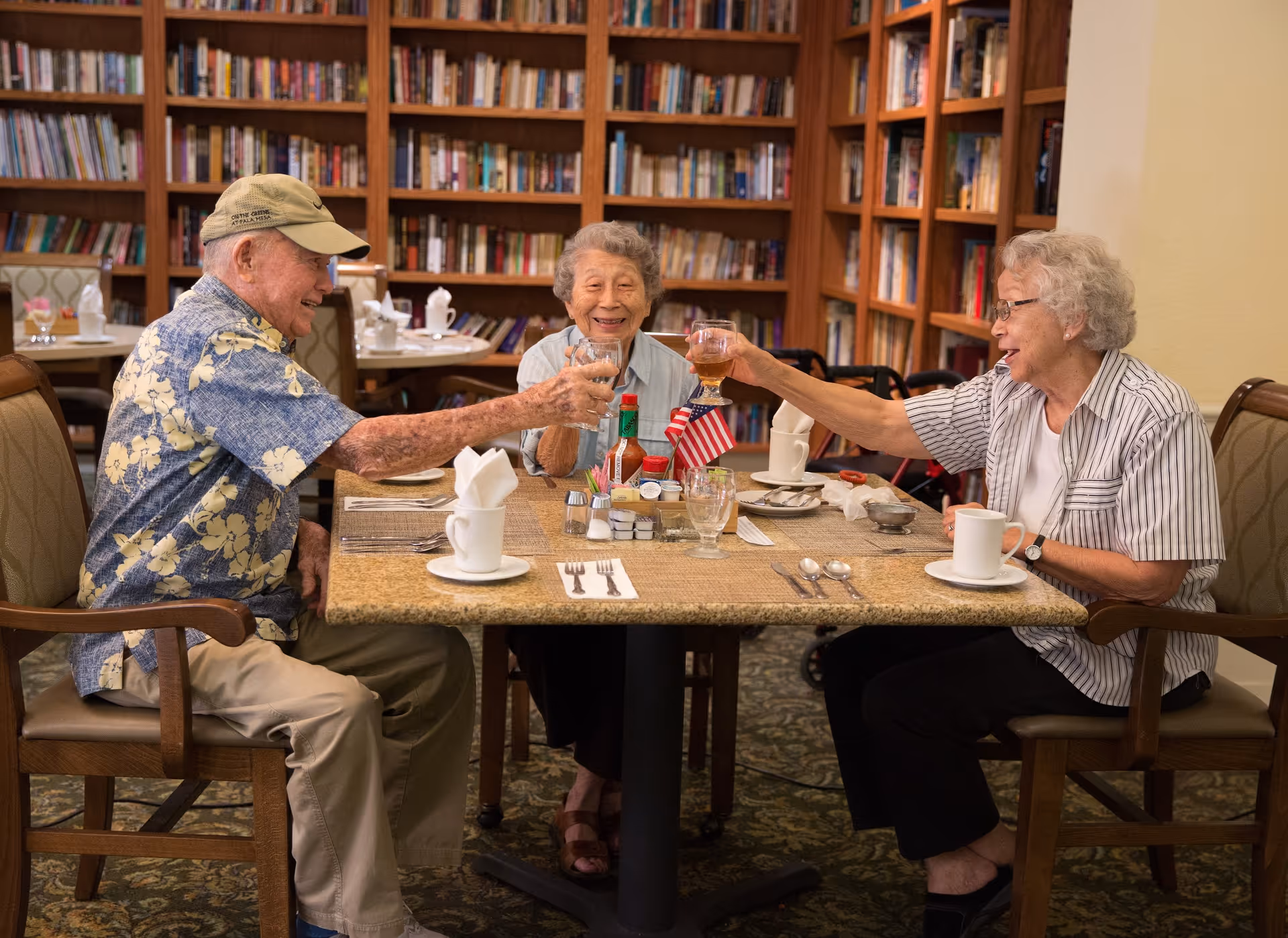 Three elderly people sitting around a dining table in a room with bookshelves, smiling and toasting with glasses. The table is set with cups, utensils, and condiments, and there is a small American flag in the center.