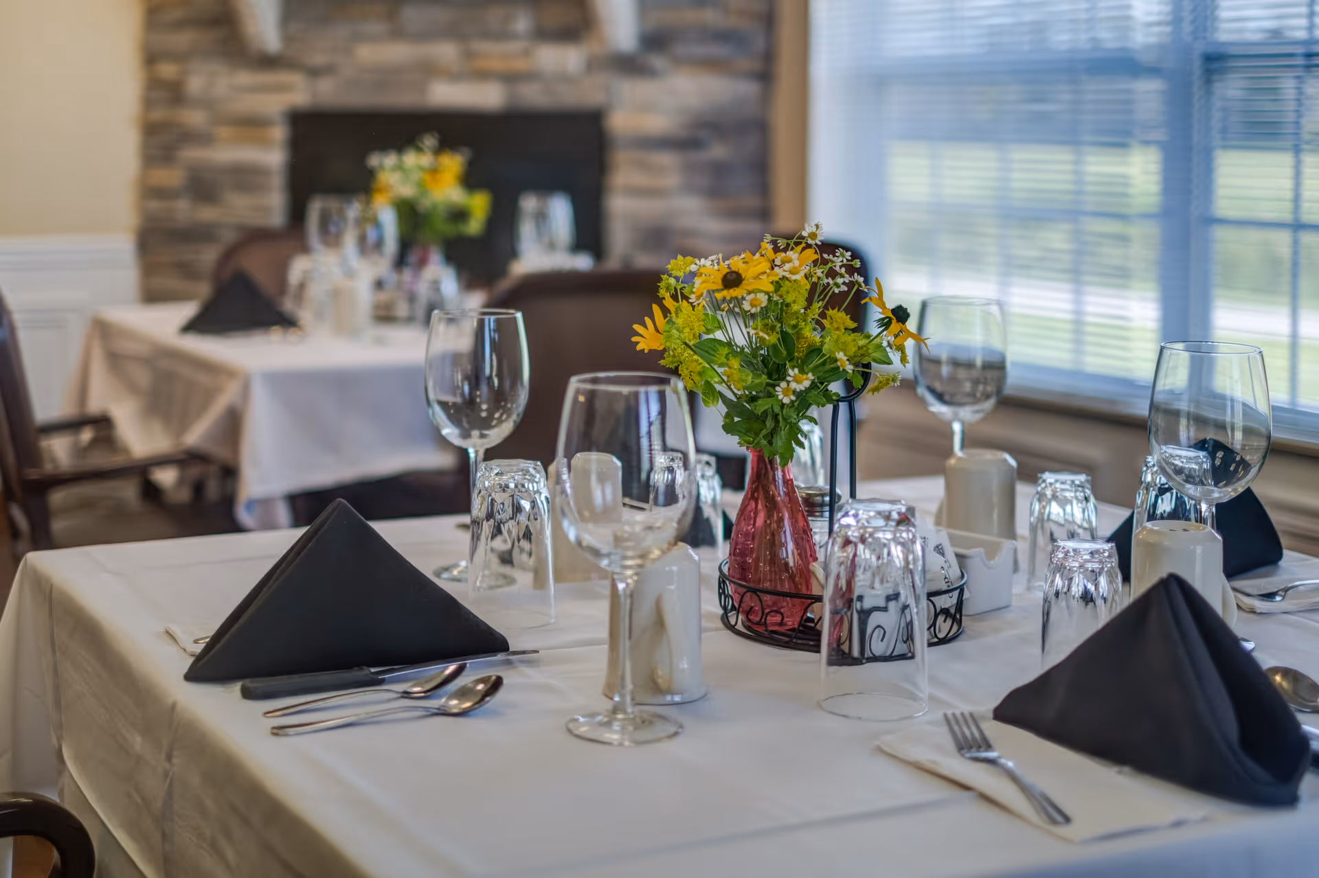 A dining table set with white tablecloth, black folded napkins, silverware, wine glasses, and a centerpiece of yellow and white flowers in a red vase. In the background, there is a stone fireplace and another similarly set table near a window with blinds.
