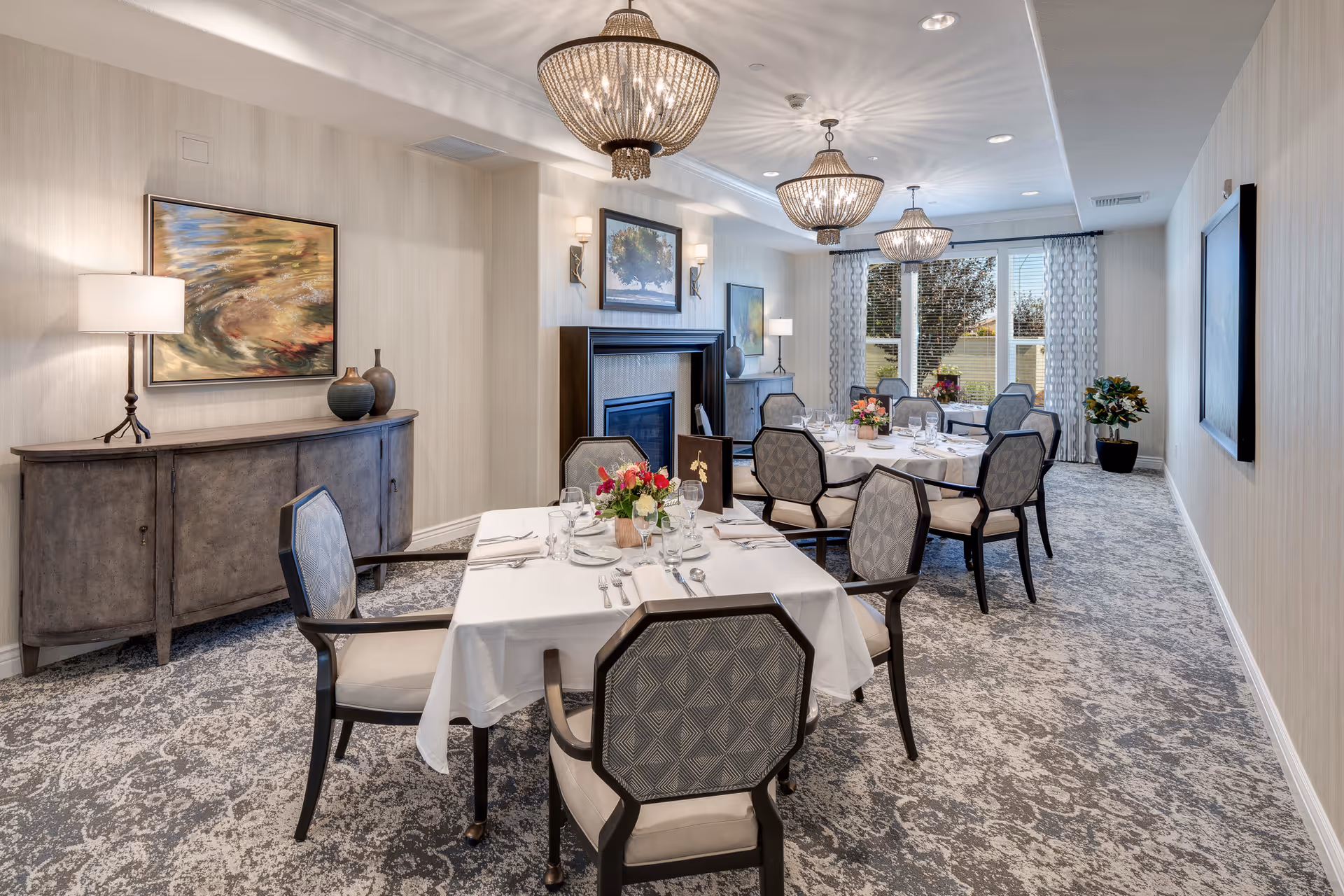 A well-lit dining room with two tables covered in white tablecloths, each set with plates, glasses, and silverware. The room features patterned carpet, elegant chairs, a sideboard with decorative vases, framed artwork on the walls, three chandeliers hanging from the ceiling, and large windows with curtains letting in natural light. There is also a fireplace with a painting above it and a potted plant near the window.
