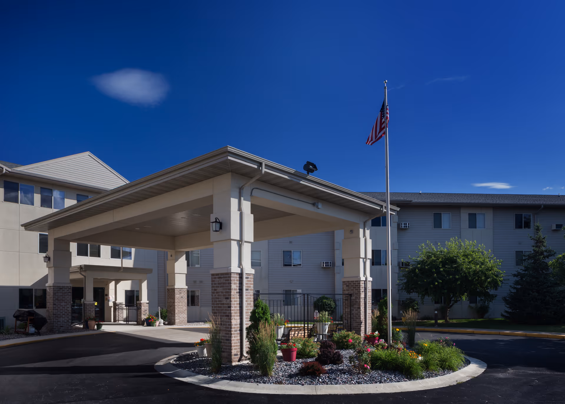 Entrance canopy and circular driveway of the Appleton Retirement Community with an American flag and landscaped flowerbed.