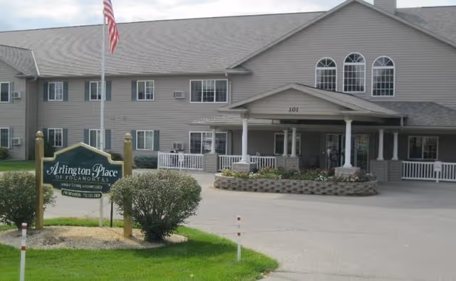 Exterior view of Arlington Place at Pocahontas senior living facility showing a two-story building with multiple windows, a covered entrance, an American flag on a flagpole, and a sign with the facility's name in front surrounded by greenery.