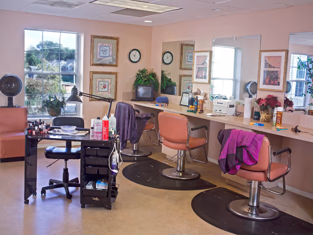 Interior view of a senior living facility salon area with three salon chairs in front of a long mirror, a small table with nail polish and manicure supplies, and a window showing greenery outside.