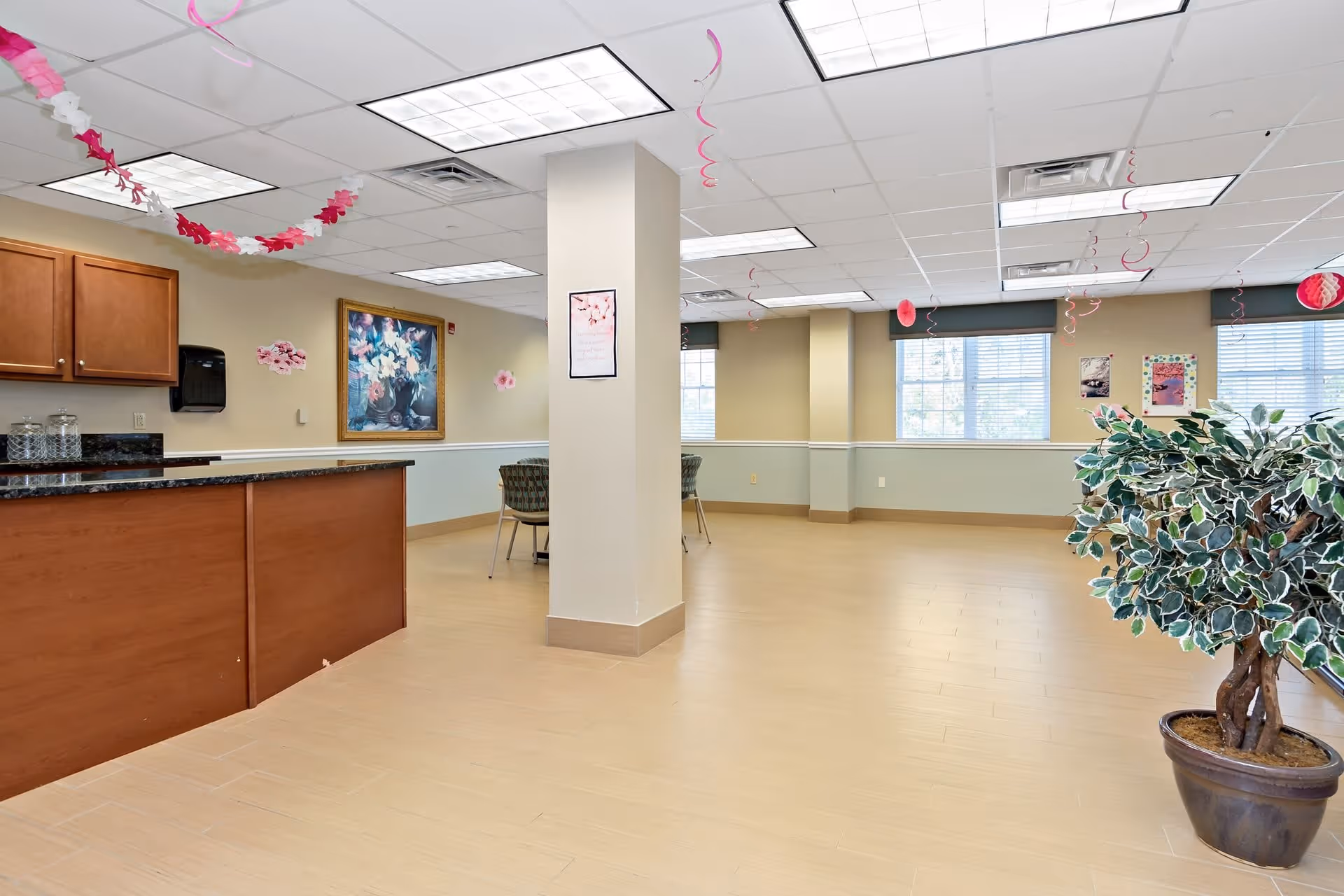 A spacious, well-lit common area with light wood flooring and beige walls. There is a wooden counter on the left side with a black granite countertop, and a few chairs and tables are visible in the background. The ceiling has fluorescent lights and pink and white decorative streamers hanging down. A large potted plant is on the right side near the windows, which have blinds partially open. Artwork and decorations adorn the walls.