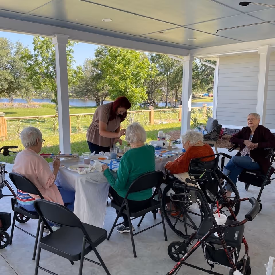 A group of elderly women sitting around a table under a covered patio, engaged in an activity with a caregiver assisting them. The setting overlooks a green lawn with trees and a body of water in the background.